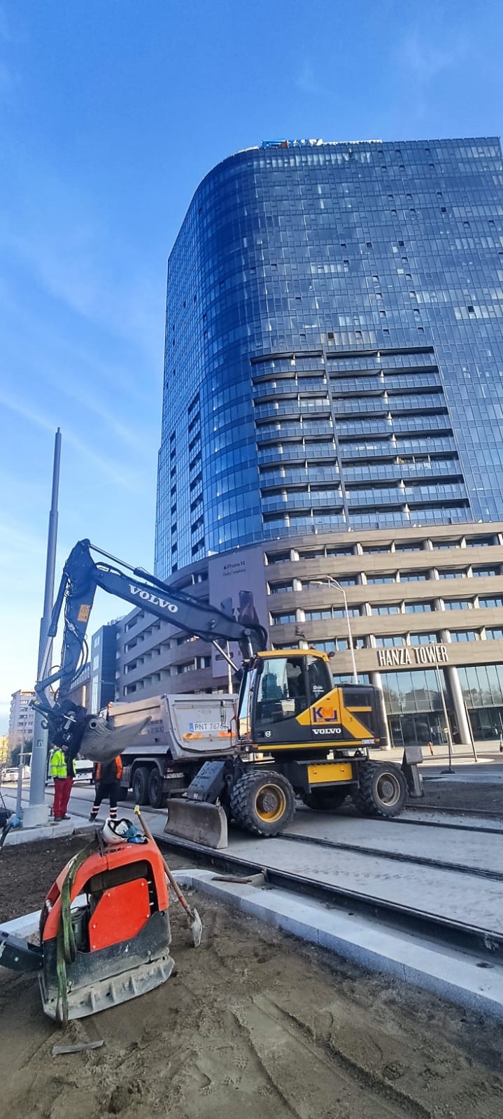Żółta koparko-ładowarka Volvo na placu budowy z ubijarką w tle, pracownicy w odblaskowych kamizelkach i wywrotka, na tle nowoczesnego budynku biurowego Hanza Tower.