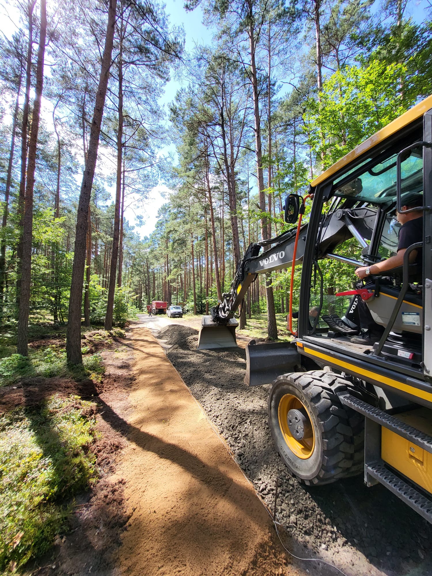 Żółta koparka Volvo wyrównuje teren leśnej drogi, widoczny operator w kabinie, w tle drzewa i samochody