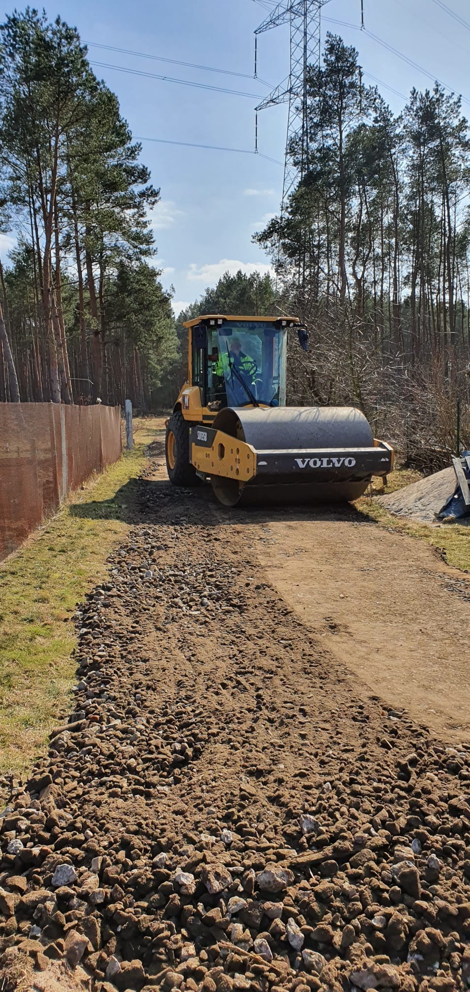 Żółty walec Volvo ubija kamienistą drogę gruntową na tle lasu i słupa wysokiego napięcia.