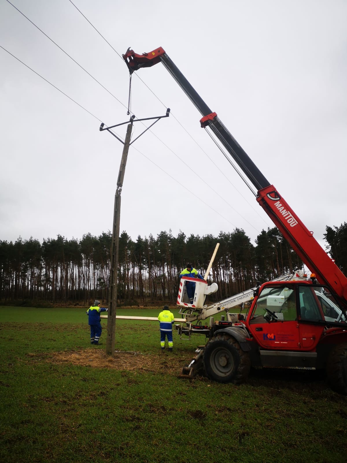 Montaż słupa energetycznego przy użyciu podnośnika koszowego Manitou na tle lasu, pracownicy w strojach roboczych.