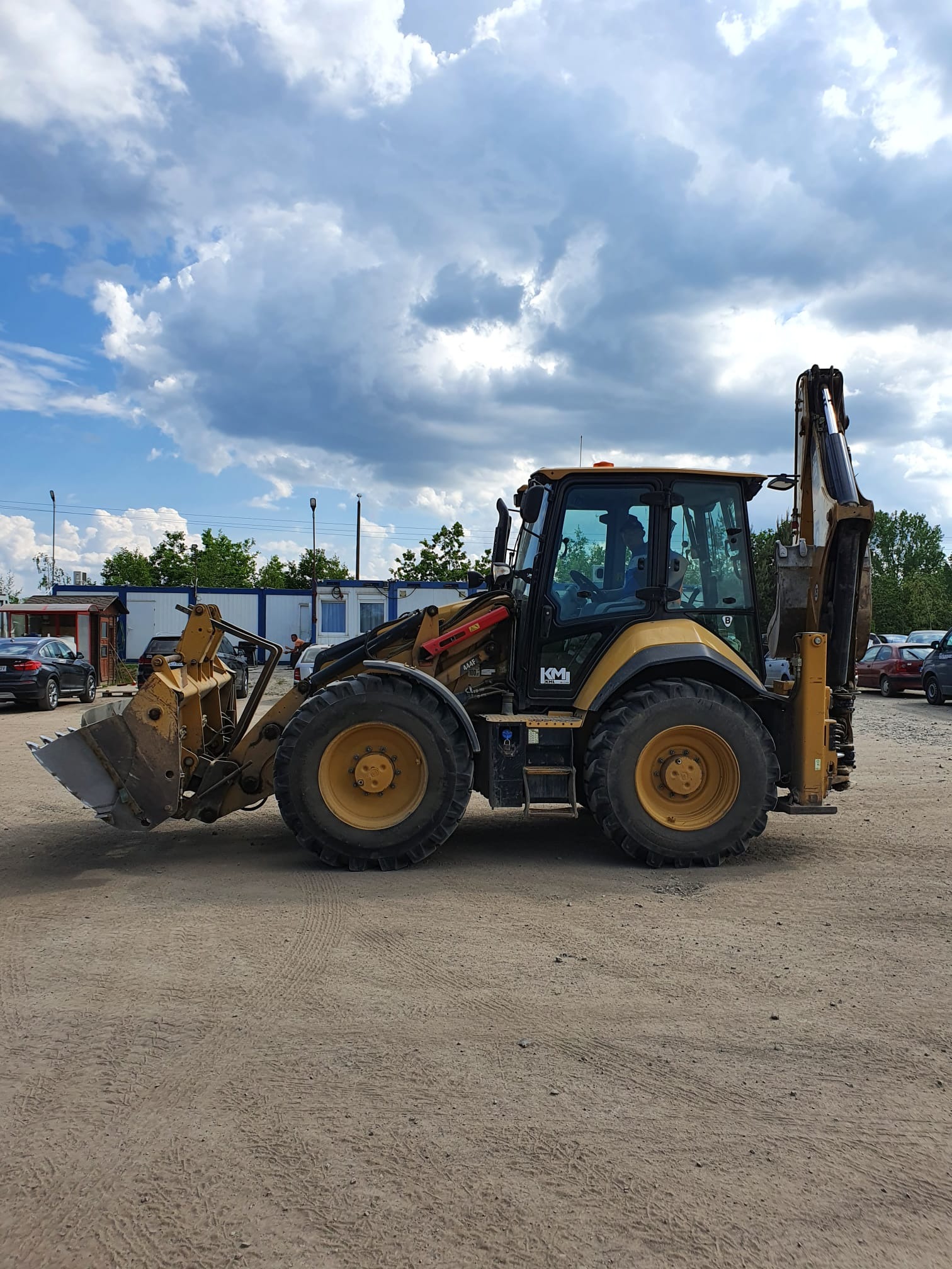 Żółto-czarna koparko-ładowarka Caterpillar na piaszczystym placu, z widocznym operatorem w kabinie, w tle biura kontenerowe i zaparkowane samochody pod błękitnym niebem z chmurami.