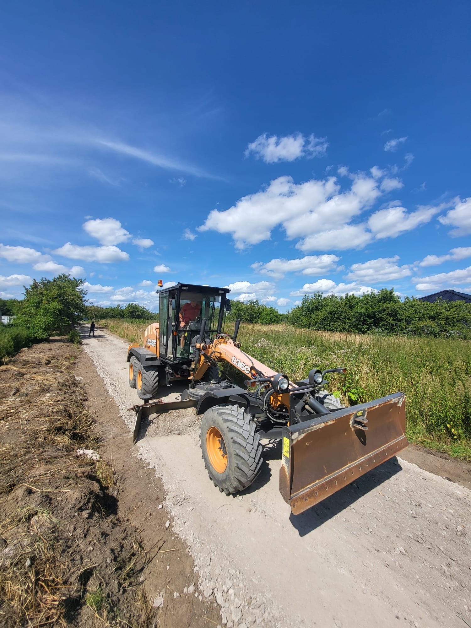 Pomarańczowa równiarka CASE 836C na drodze gruntowej z widocznym operatorem w kabinie, świeżo wyrównany teren po prawej stronie, błękitne niebo z obłokami w tle.