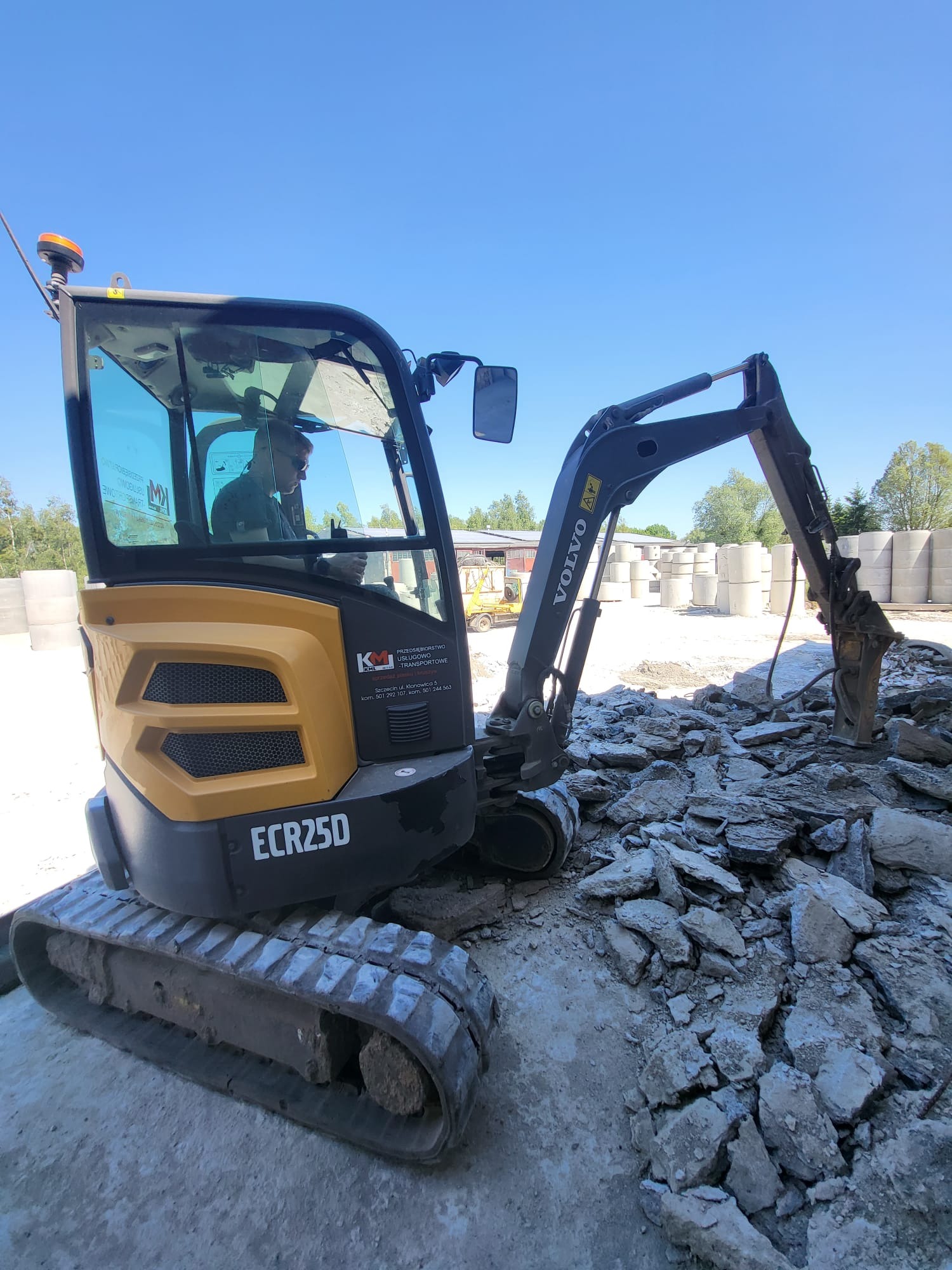 Żółto-czarna minikoparka Volvo ECR25D z operatorem w kabinie, wyposażona w młot hydrauliczny, kruszy gruz na betonowej powierzchni; w tle widoczne betonowe kręgi.