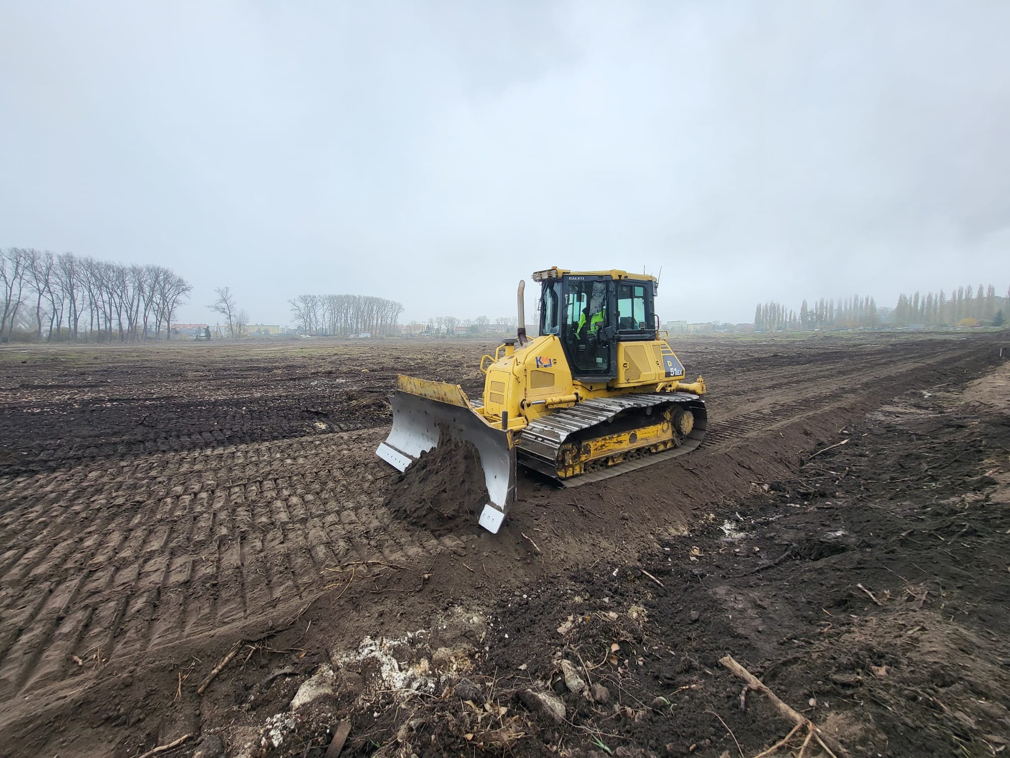 Żółty spychacz Caterpillar D51EX na rozmokłym, błotnistym polu, wyrównujący teren, widoczny operator w odblaskowej kamizelce, pochmurne niebo w tle.