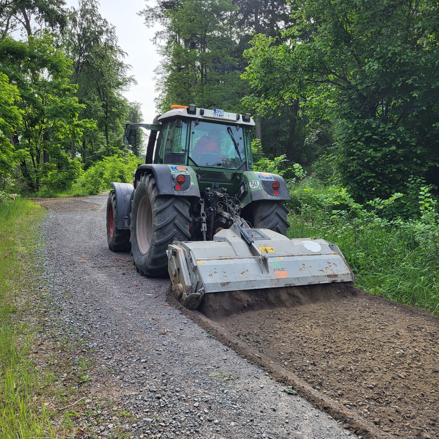 Traktor z frezem wyrównuje szutrową drogę w Dołujach, otoczoną zielenią lasu.