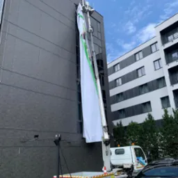 Podnośnik koszowy z długim, białym banerem reklamowym z zielonym obramowaniem, umieszczonym na elewacji budynku w nowoczesnym stylu.