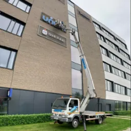 Pracownik w koszu podnośnika na tle biurowca z logo 'Bank Polski' i 'Uniqa', prawdopodobnie wykonuje prace konserwacyjne lub montażowe.