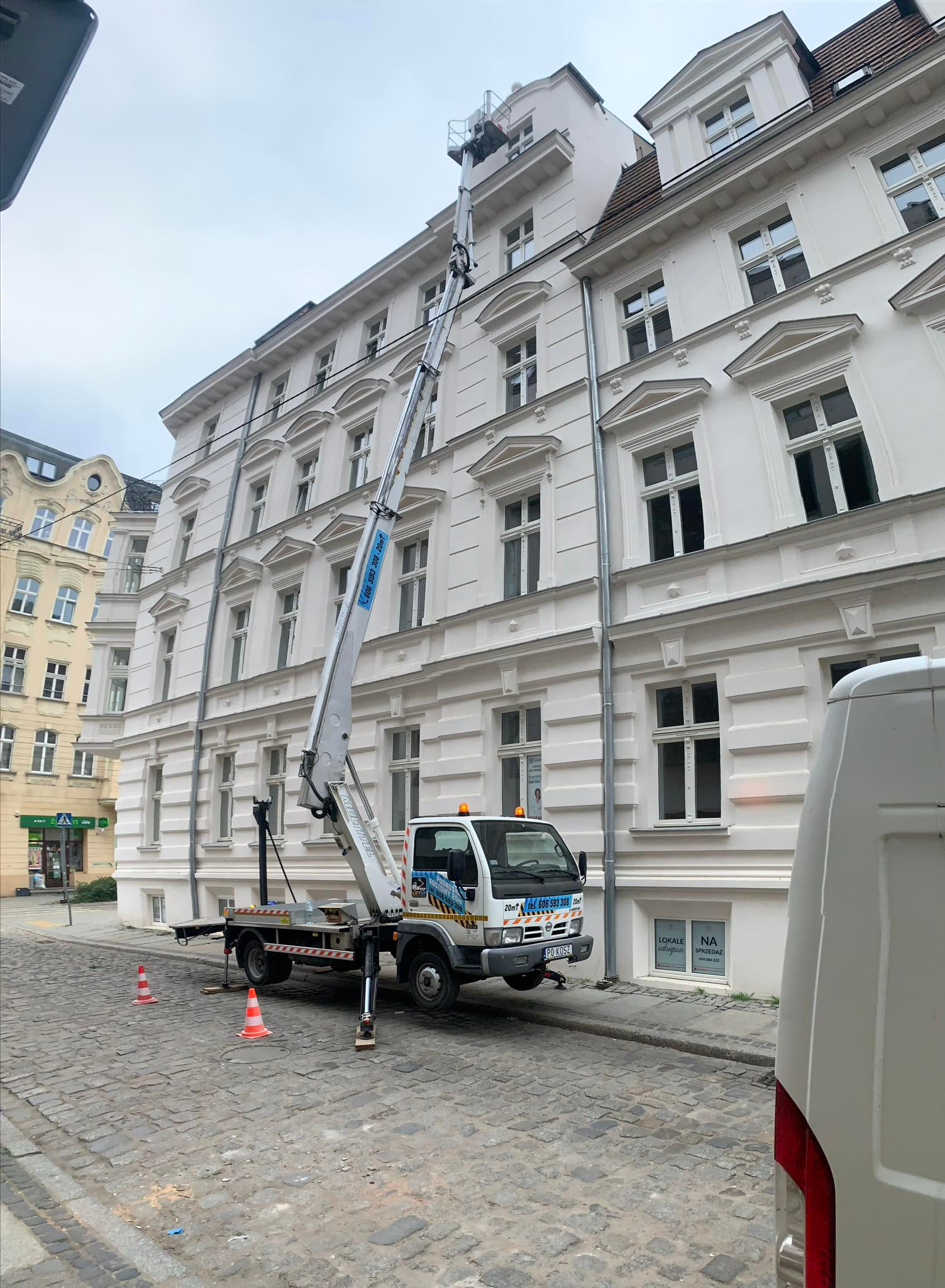 Platforma robocza na wysięgniku podnośnika koszowego sięga okna kamienicy w Poznaniu. Na brukowanej ulicy ustawione pachołki ostrzegawcze, częściowo widoczny biały samochód dostawczy.