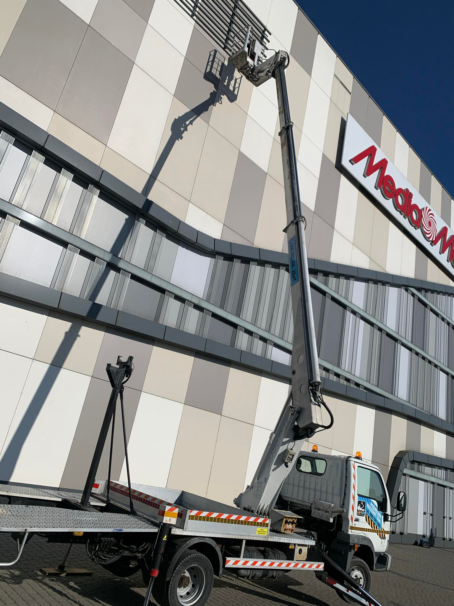 Podnośnik koszowy na pojeździe, z wysięgnikiem ustawionym przy elewacji budynku z logo Media Markt, widoczne detale konstrukcyjne elewacji.