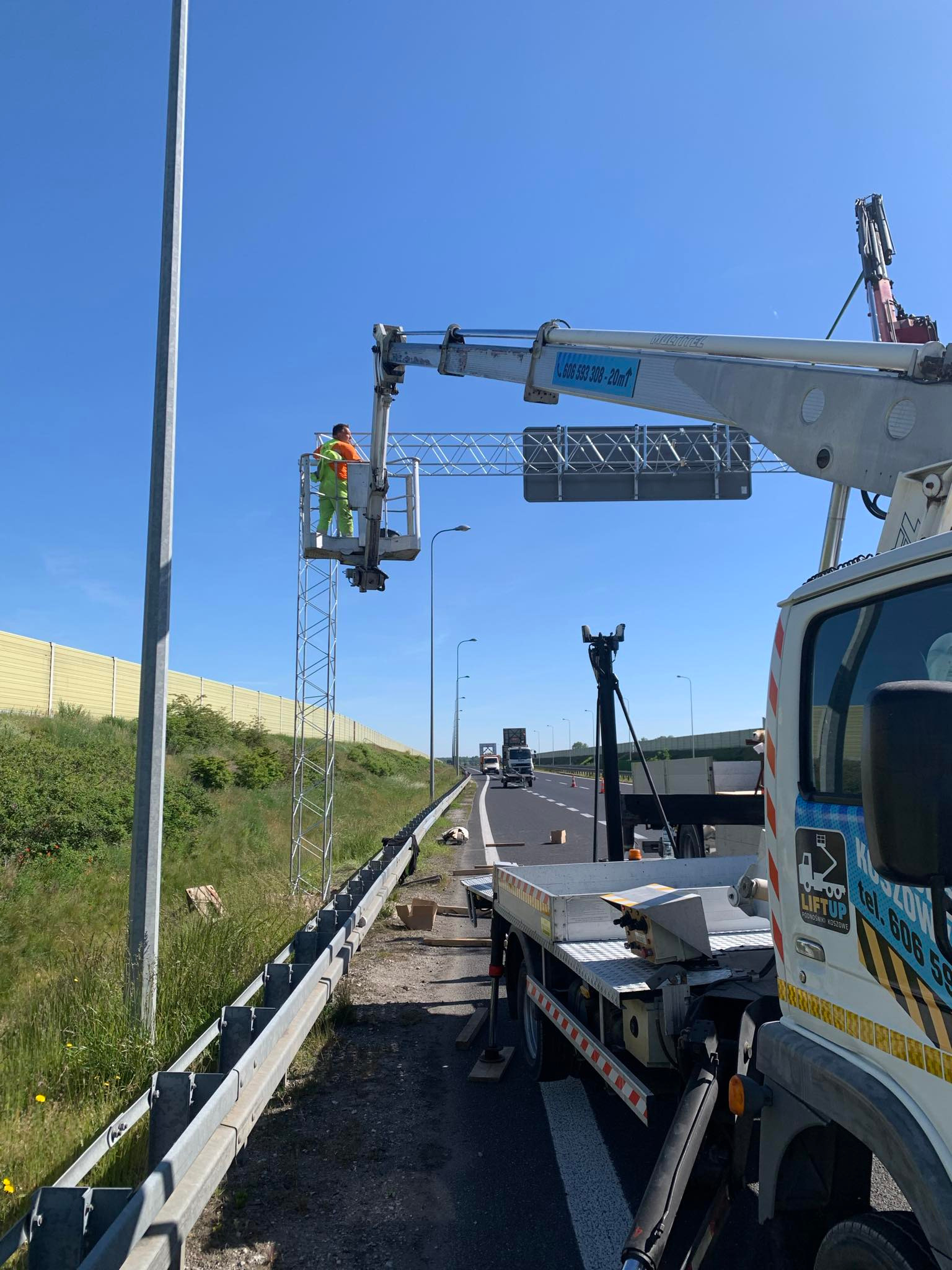 Pracownik w pomarańczowej kamizelce i zielonym kombinezonie w koszu podnośnika na autostradzie, instaluje lub naprawia metalową konstrukcję nad jezdnią, widoczny samochód z podnośnikiem koszowym.