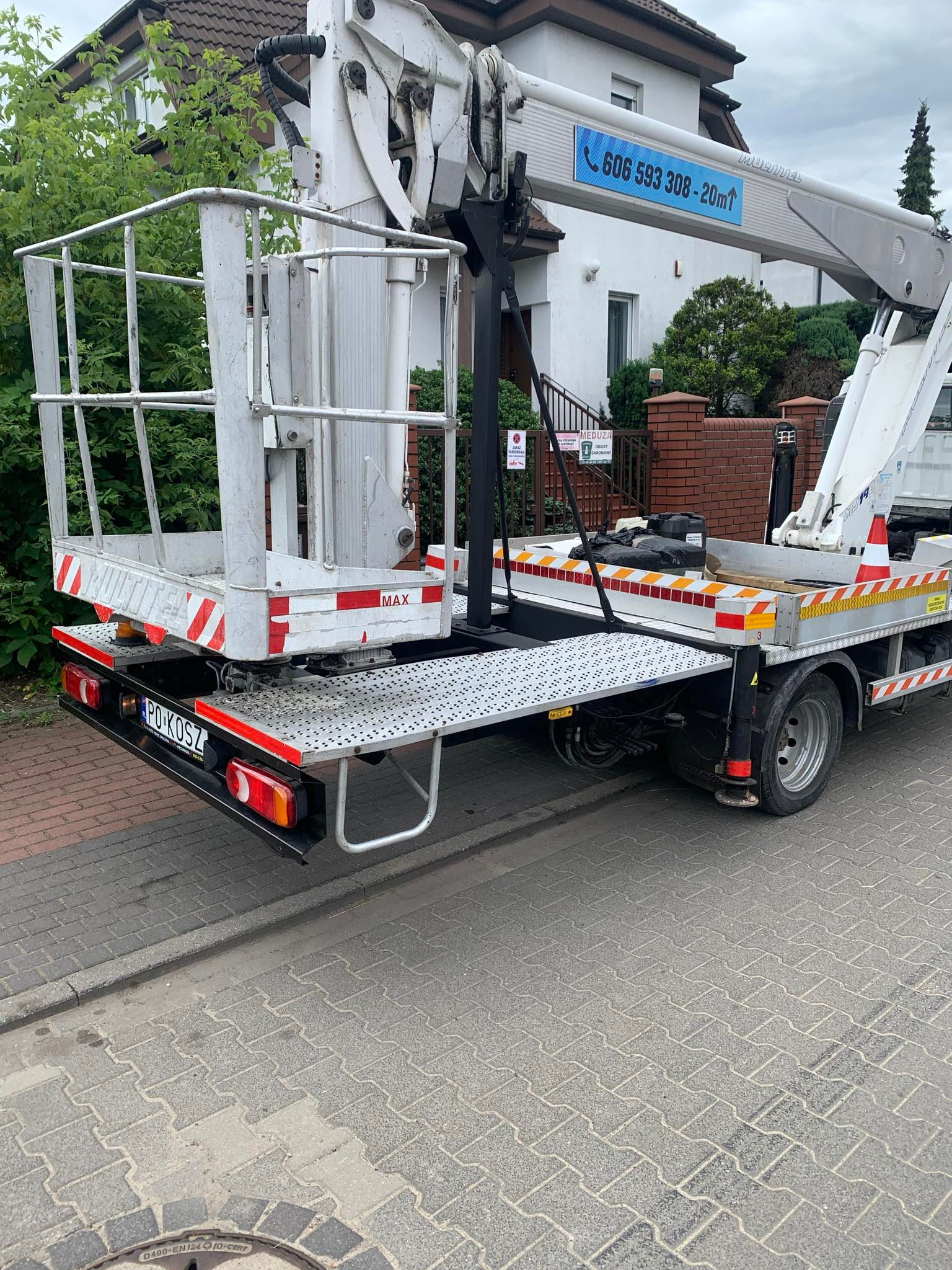Biały podnośnik koszowy na samochodzie, widoczny z tyłu, z podniesionym koszem i numerem telefonu kontaktowym na wysięgniku, zaparkowany na chodniku z kostki brukowej przed budynkiem mieszkalnym.