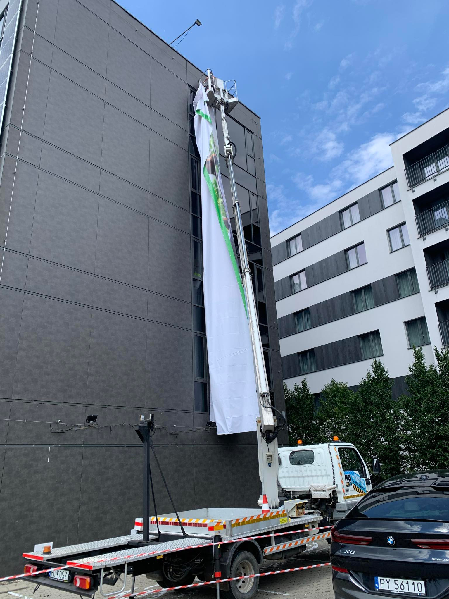 Podnośnik koszowy z długim, białym banerem reklamowym z zielonym obramowaniem, umieszczonym na elewacji budynku w nowoczesnym stylu.