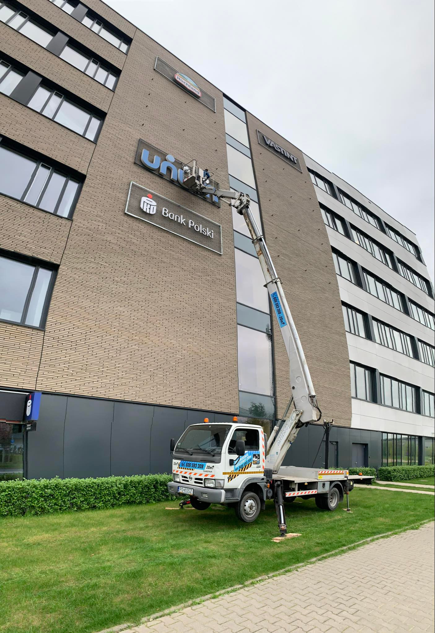 Pracownik w koszu podnośnika na tle biurowca z logo 'Bank Polski' i 'Uniqa', prawdopodobnie wykonuje prace konserwacyjne lub montażowe.