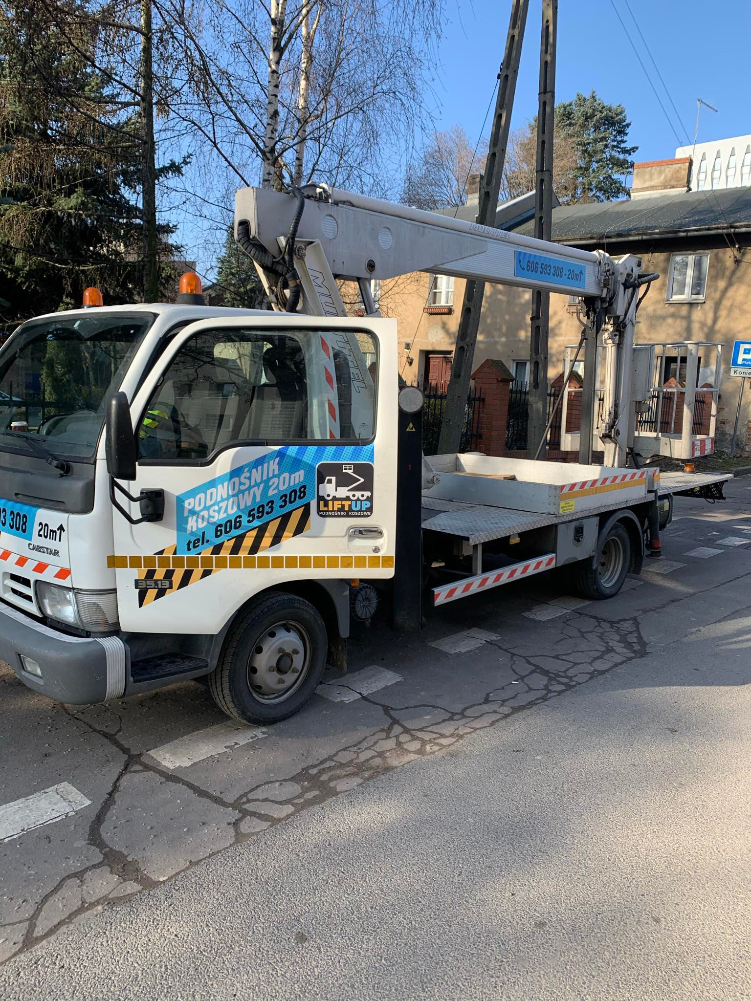 Biała ciężarówka z podnośnikiem koszowym zaparkowana na pasach dla pieszych, z widocznym logo firmy i numerem telefonu na boku, na tle budynków i drzew.