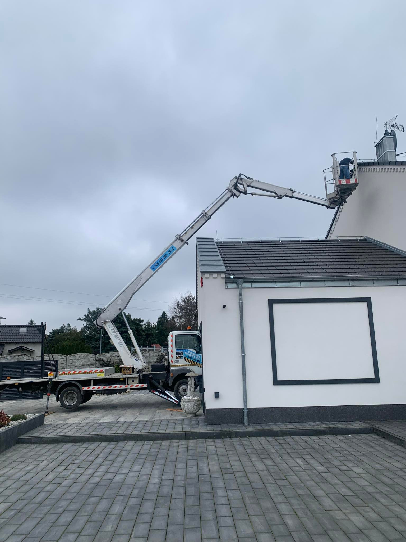 Pracownik w koszu podnośnika teleskopowego, na tle białej elewacji budynku z szarą ramą dekoracyjną, wykonuje prace na wysokości przy dachu. W tle widoczny zaparkowany samochód z podnośnikiem.