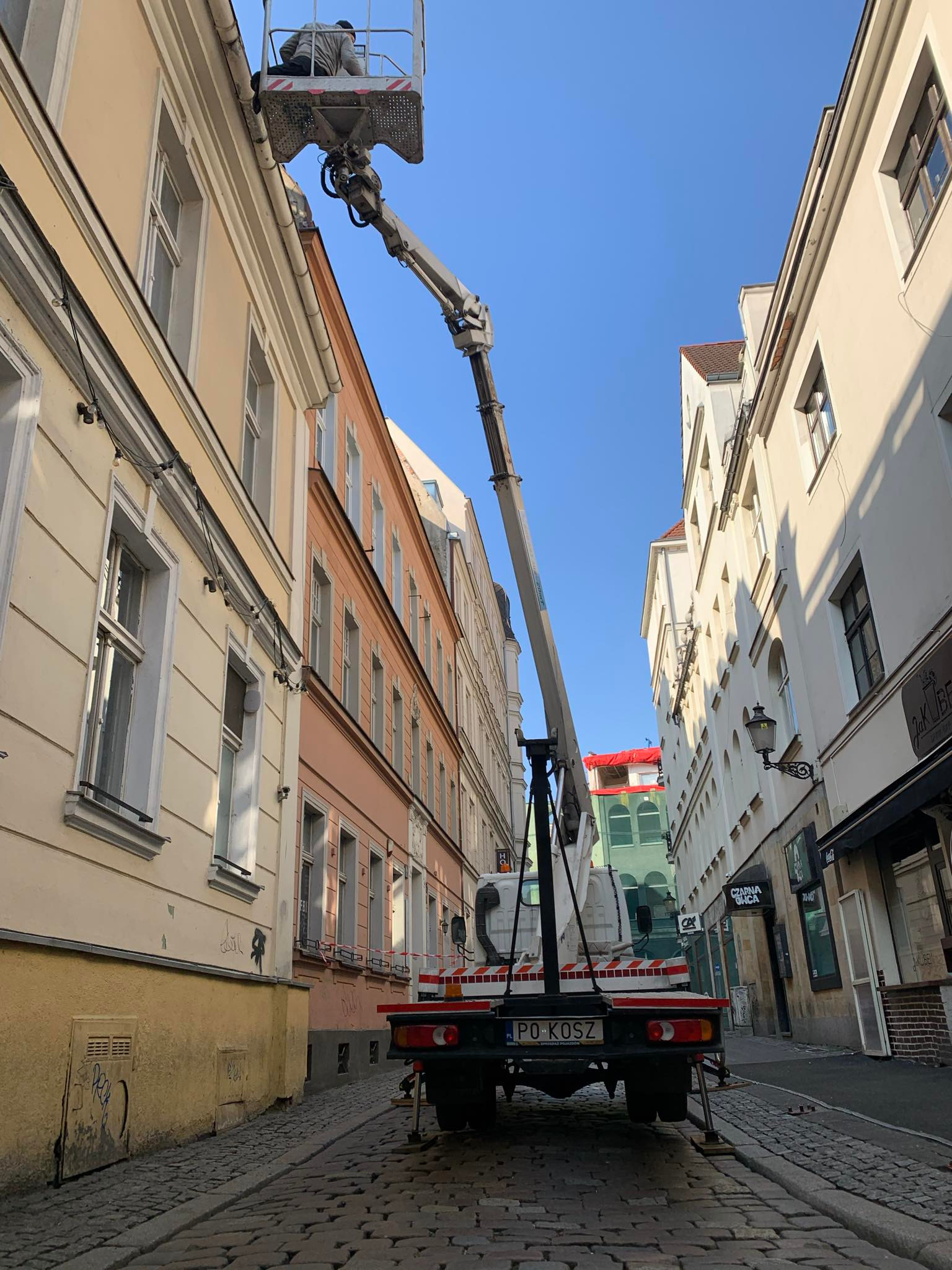 Pracownik w koszu podnośnika teleskopowego na tle kamienicy, prawdopodobnie wykonuje prace konserwacyjne na wysokości w wąskiej uliczce miejskiej.