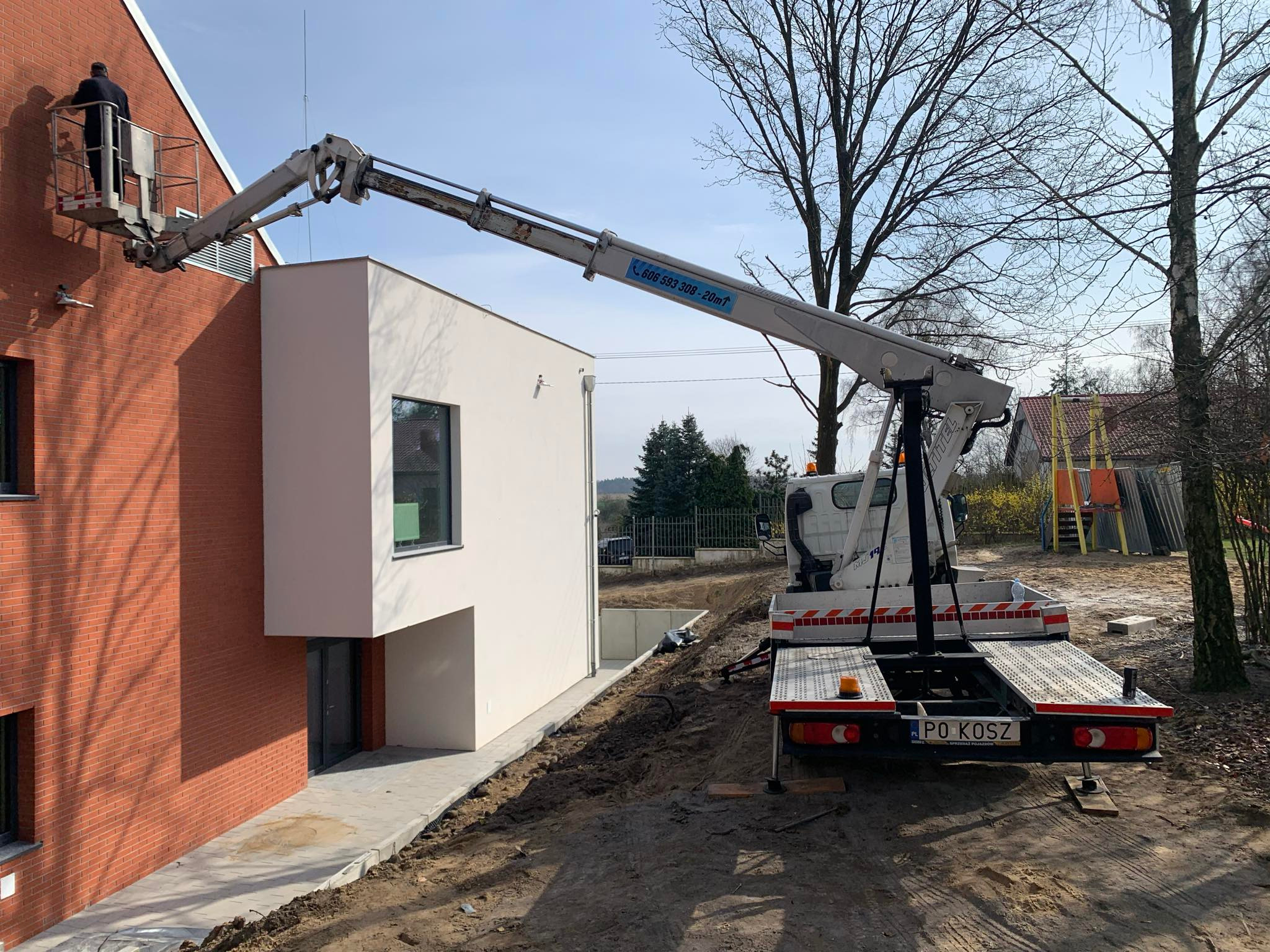 Pracownik w koszu podnośnika teleskopowego przy dwukondygnacyjnym budynku z ceglaną elewacją, białą dobudówką i widocznymi elementami instalacji na ścianie.
