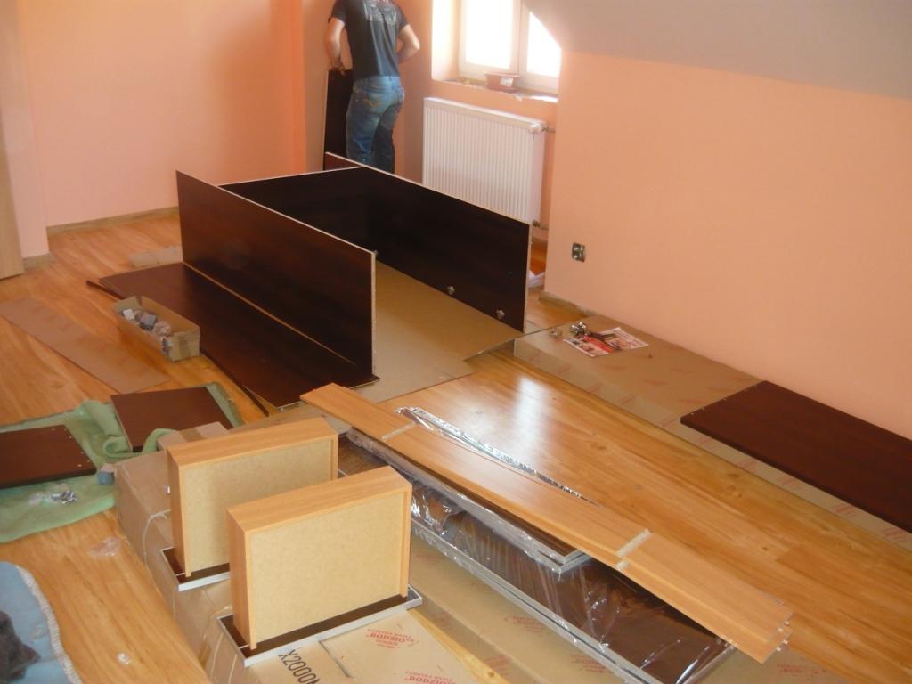 Montaż szafy: widok na rozłożone elementy meblowe z ciemnego drewna na podłodze w pokoju, w tle osoba montująca kolejny element.