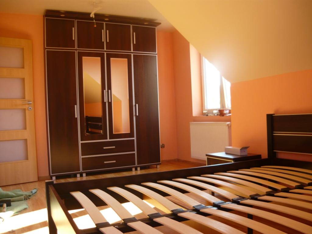 Ciemnobrązowa szafa z lustrami i jasnymi obramowaniami, dopasowana do skośnego sufitu w pomarańczowym pokoju z łóżkiem w trakcie montażu.