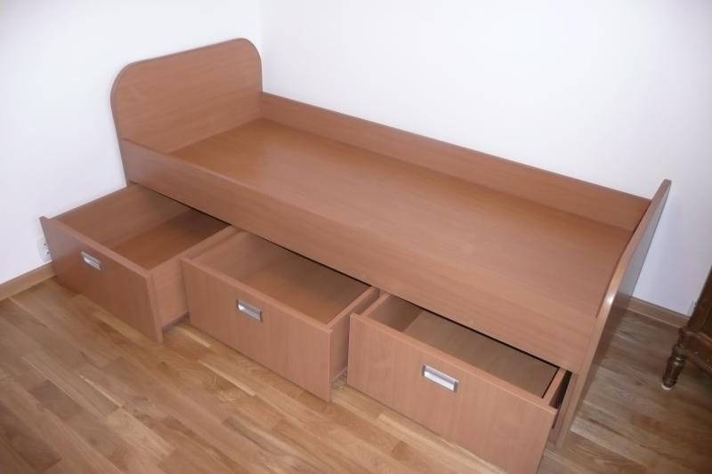 Łóżko jednoosobowe z trzema wysuwanymi szufladami w kolorze jasnego drewna, widoczne na parkiecie, dwie szuflady otwarte.
