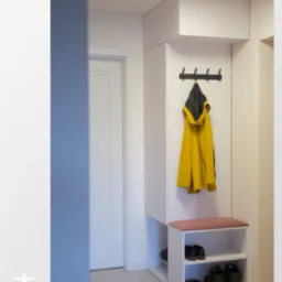 Biała szafa wnękowa z siedziskiem i wieszakami na ubrania w przedpokoju. Na półkach widoczne buty. Żółta kurtka wisi na wieszaku.