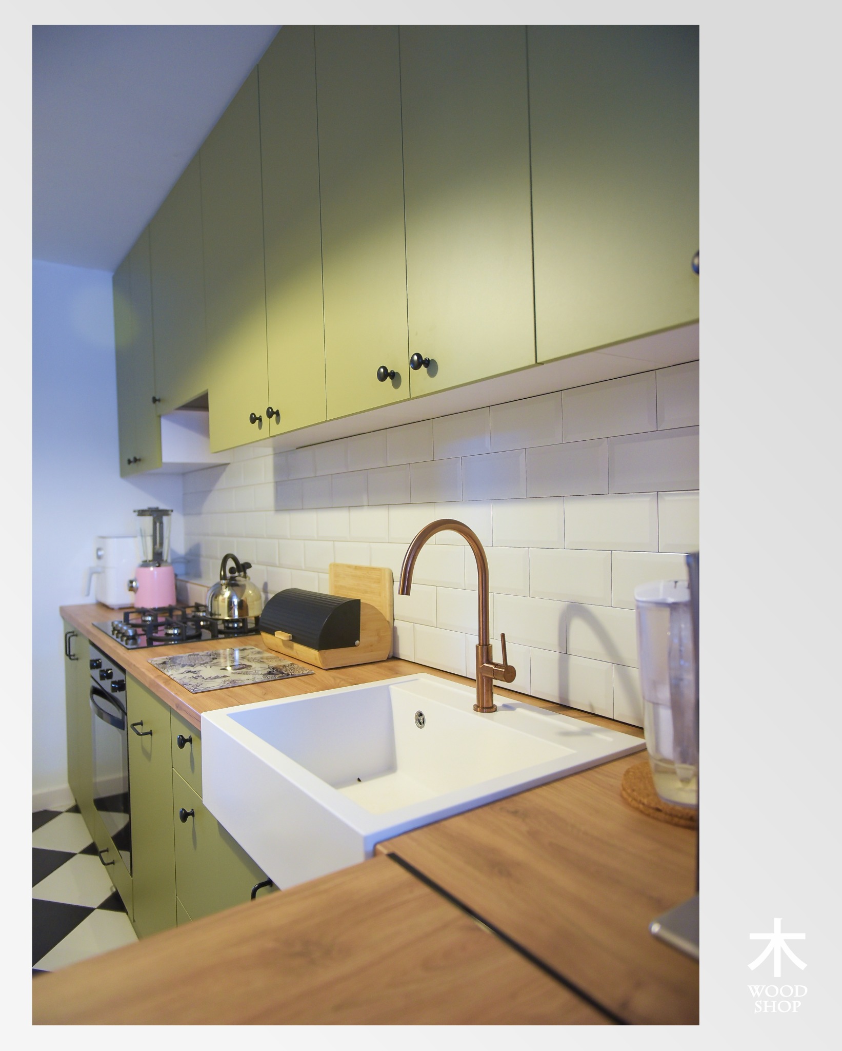 Ustawna kuchnia z zielonymi szafkami, białymi płytkami i drewnianym blatem. Miedziana bateria nad białym zlewem farmerskim. Na blacie czajnik i toster.
