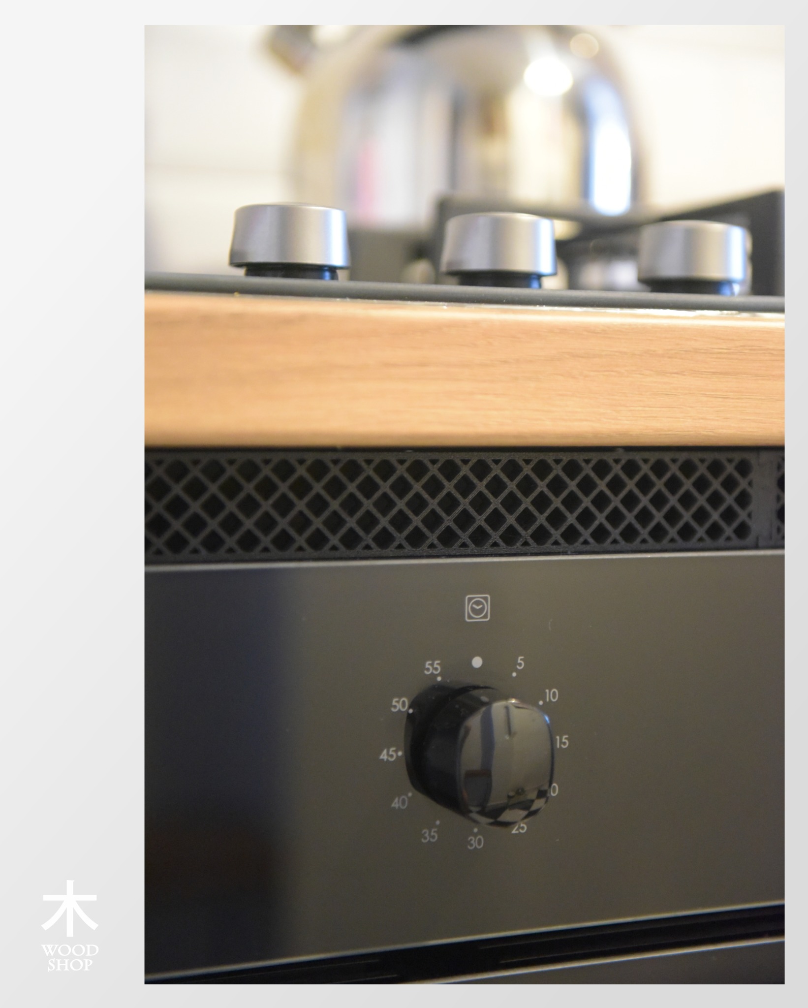 Fragment zabudowy kuchennej: detale płyty gazowej z drewnianym blatem i piekarnika z pokrętłem. Widoczny srebrny czajnik w tle.