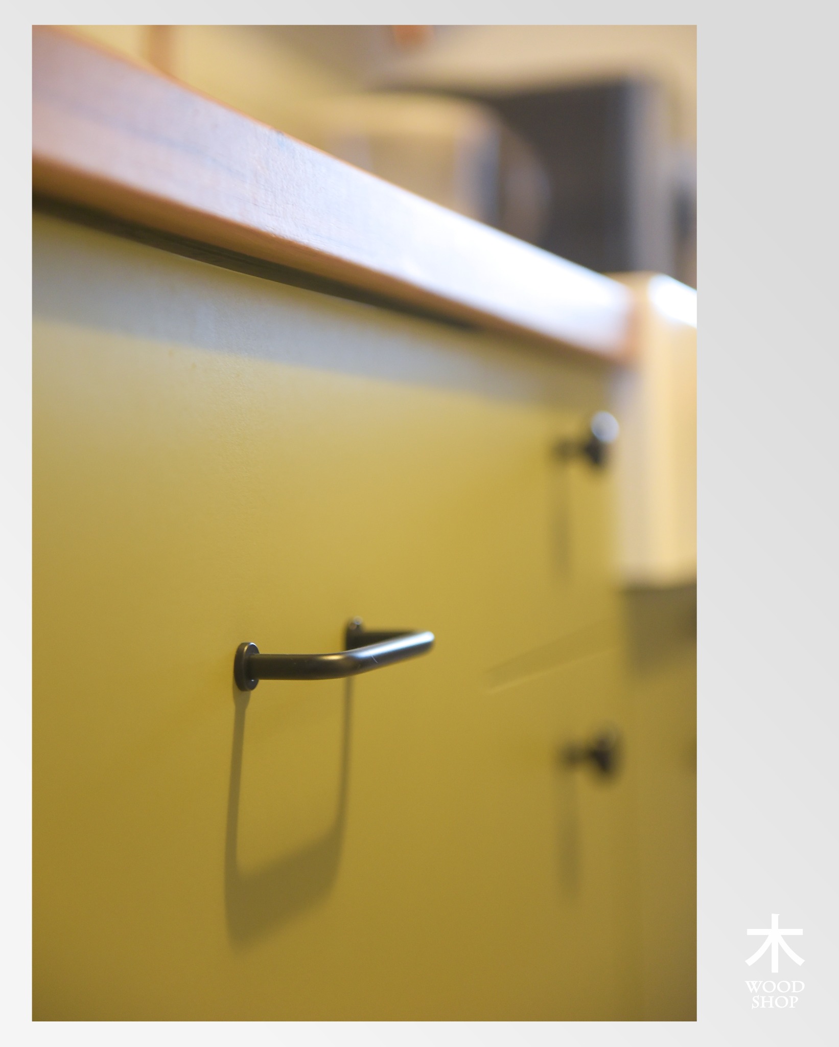 Żółta szafka z drewnianym blatem i czarnym uchwytem, widok z bliska. Widoczne logo firmy w prawym dolnym rogu.