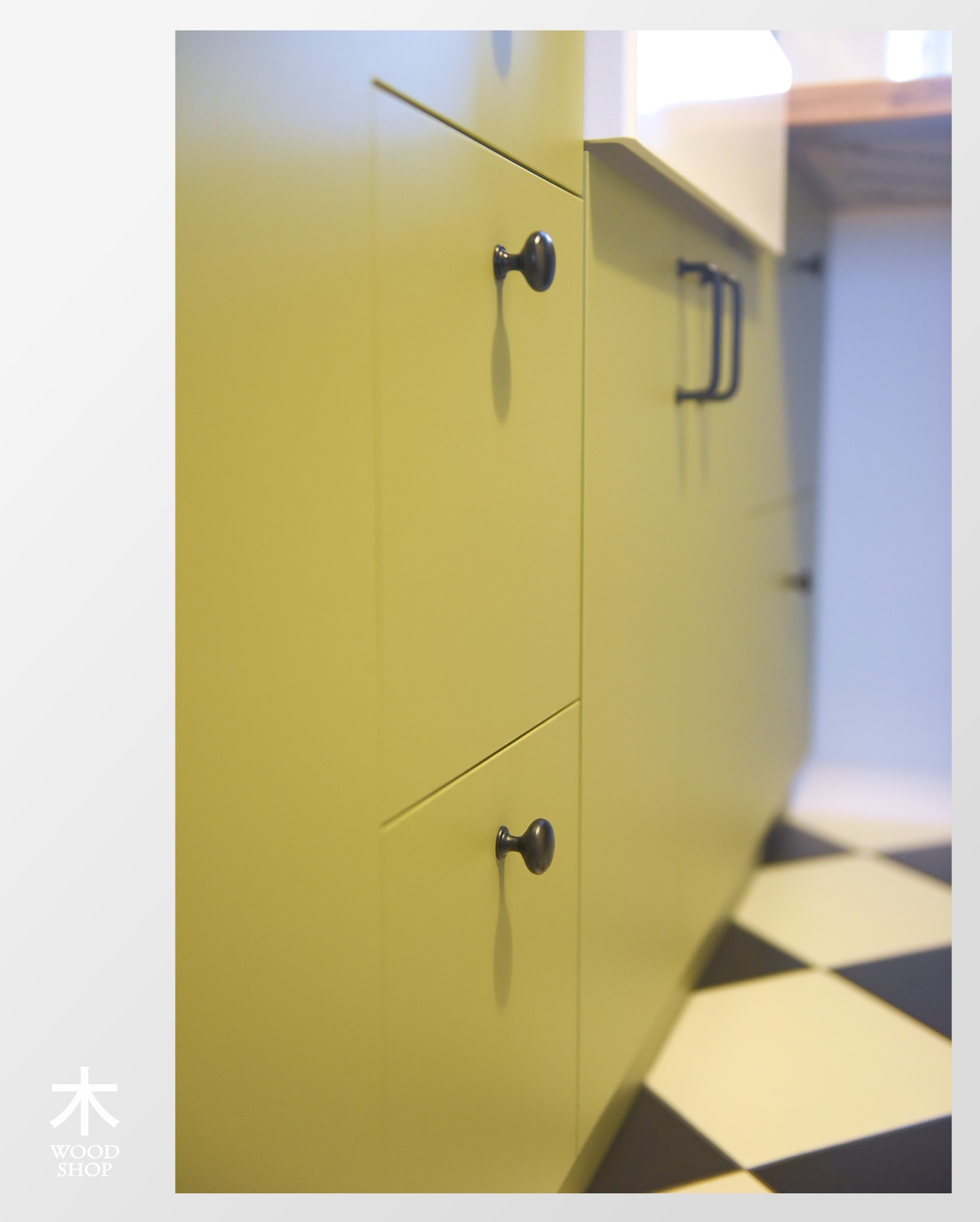 Fragment zabudowy kuchennej z oliwkowymi frontami i czarnymi, okrągłymi uchwytami. Ujęcie z dołu, widoczna szachownica na podłodze i fragment blatu.