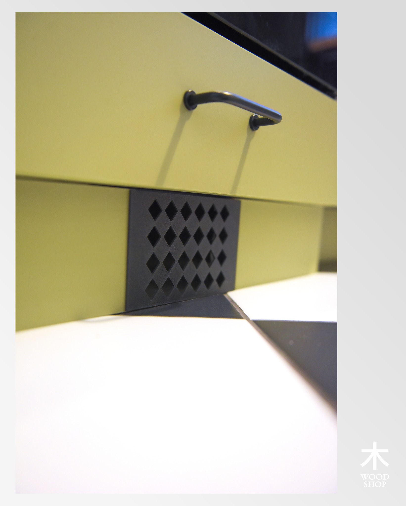 Żółta szafka z czarnym uchwytem i kratką wentylacyjną na tle biało-czarnej podłogi. Detal wykonania mebla na wymiar.