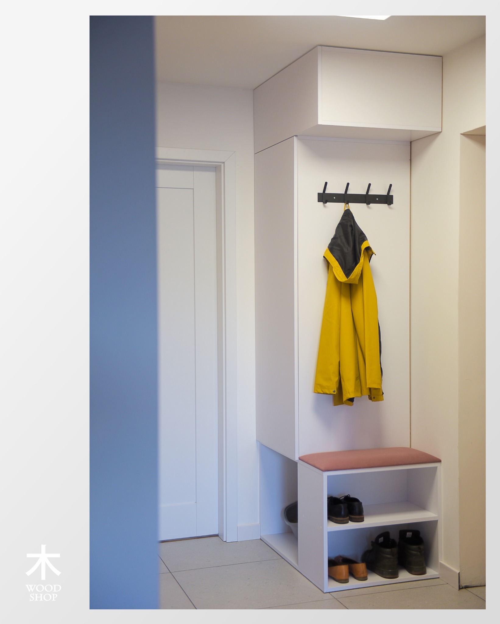 Biała szafa wnękowa z siedziskiem i wieszakami na ubrania w przedpokoju. Na półkach widoczne buty. Żółta kurtka wisi na wieszaku.