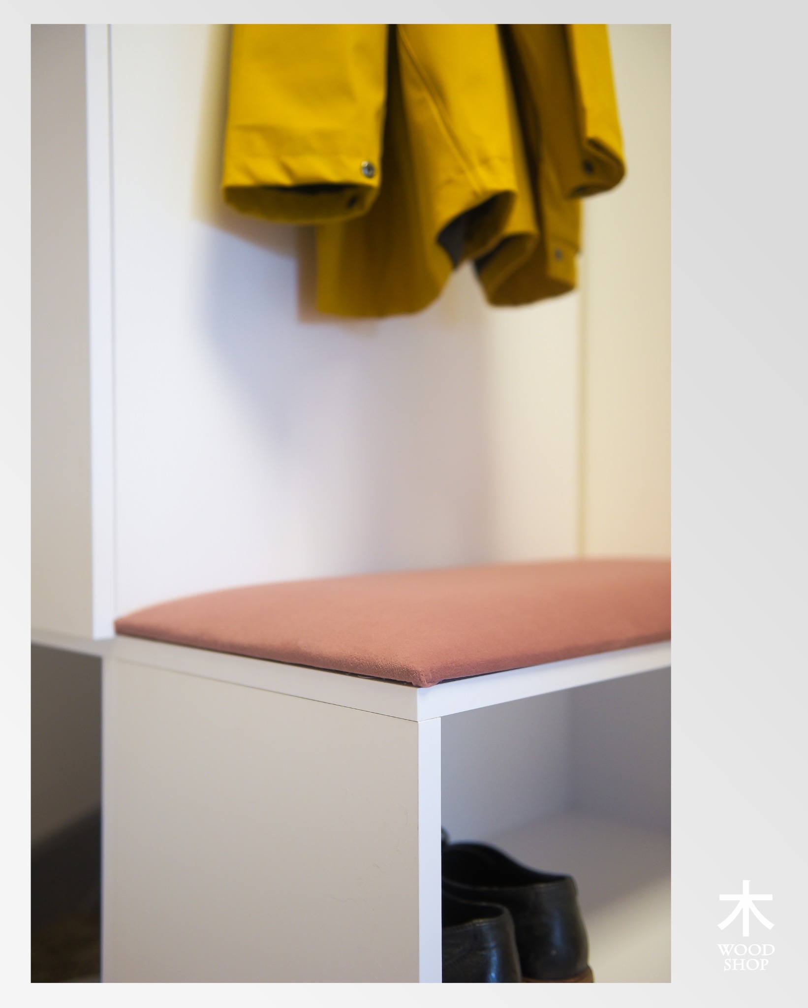 Biała szafka z siedziskiem w odcieniu brudnego różu i wiszącą żółtą kurtką. W dolnej części widoczne buty. Minimalistyczny design.