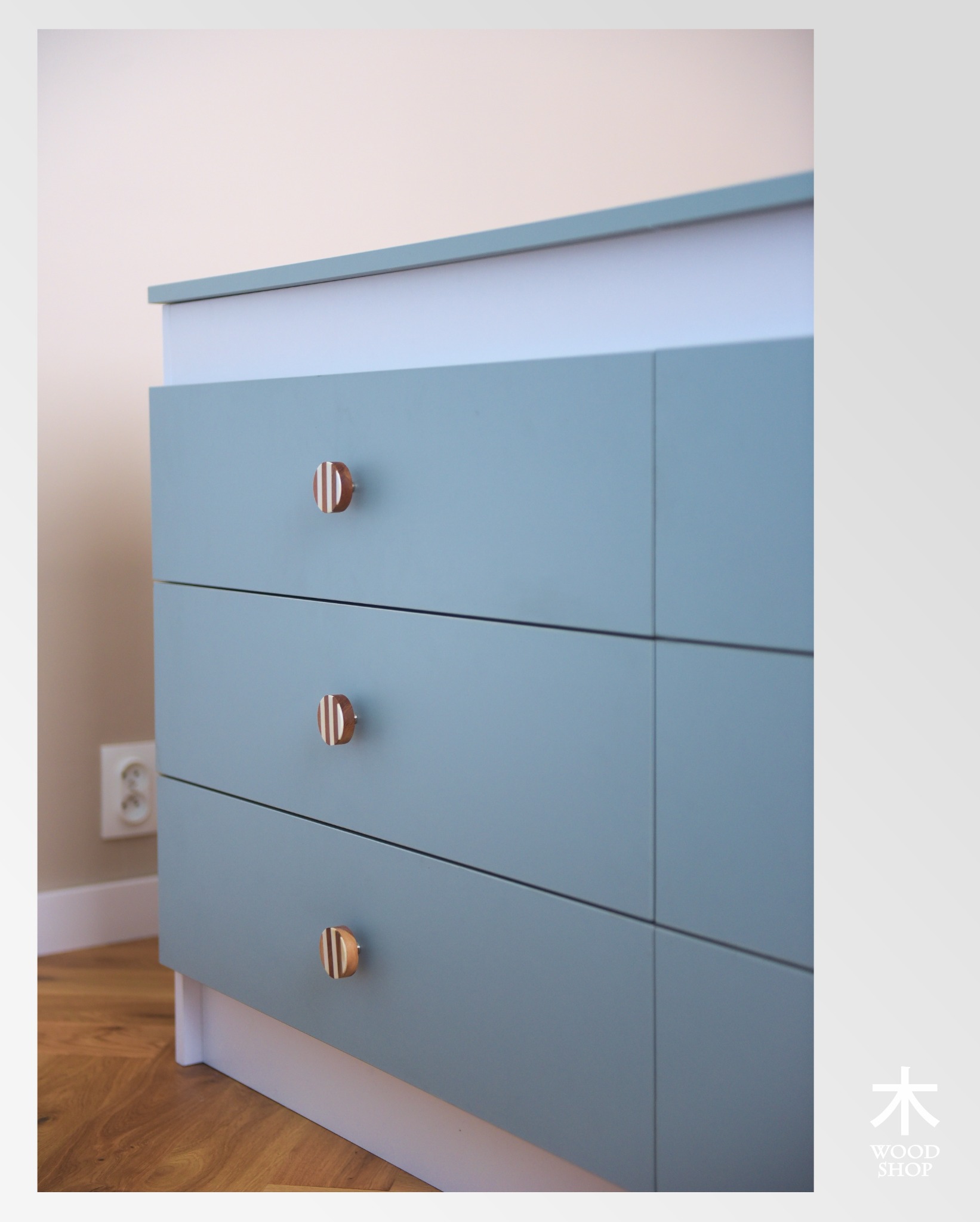 Niebieska komoda z trzema szufladami i okrągłymi, drewnianymi uchwytami w paski, na tle jasnej ściany i drewnianej podłogi. Widoczny fragment gniazdka elektrycznego.