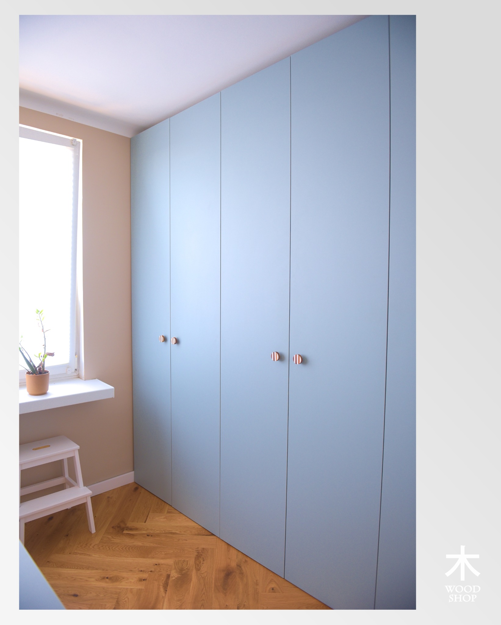 Błękitna szafa wnękowa z dyskretnymi uchwytami na tle parkietu w jodełkę. Obok okno z rośliną w doniczce i biały taboret.
