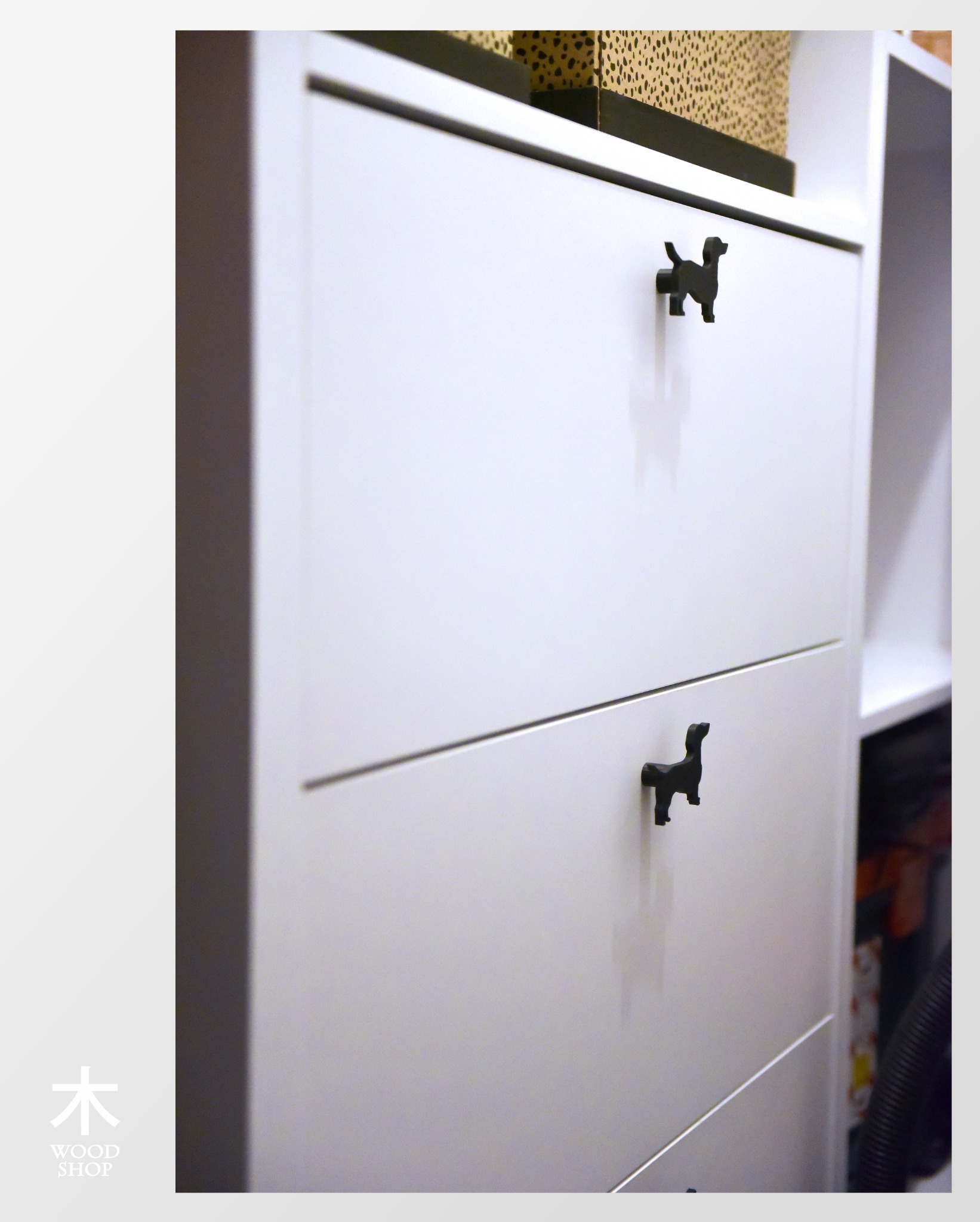 Biała szafa z nietypowymi uchwytami w kształcie psów, widoczne dwie szuflady, na górze pudełko w panterkę, minimalistyczny design.