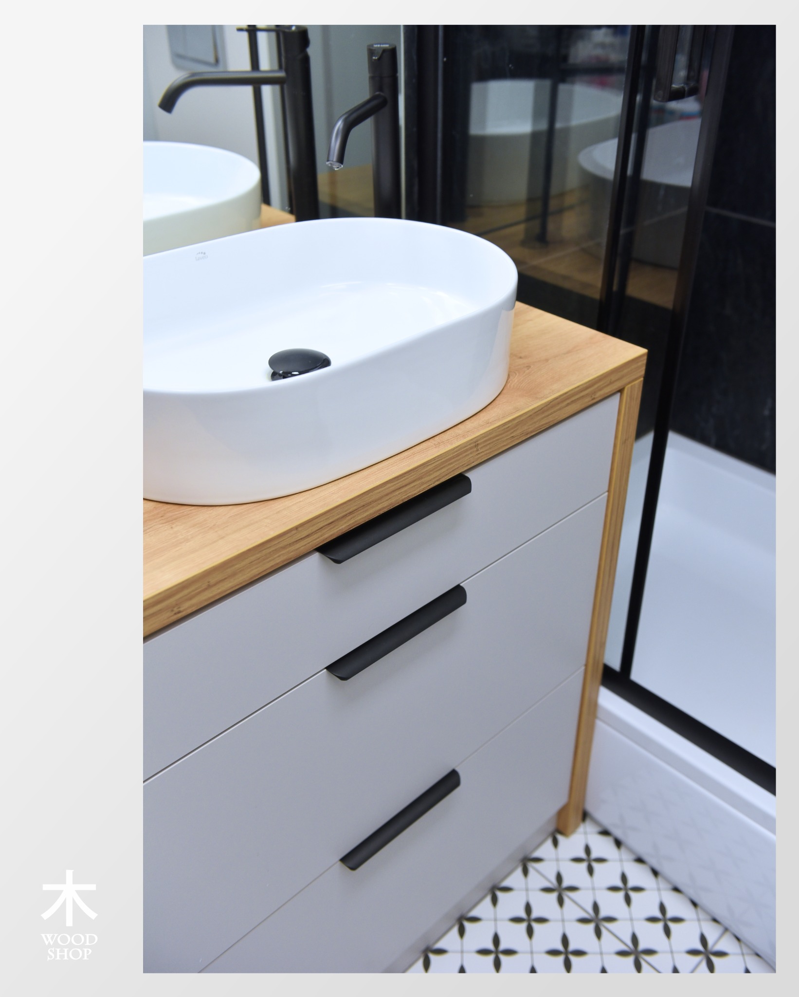 Nowoczesna szafka łazienkowa z szufladami, blat z jasnego drewna, biała umywalka nablatowa, czarne, minimalistyczne uchwyty i armatura. W tle kabina prysznicowa.