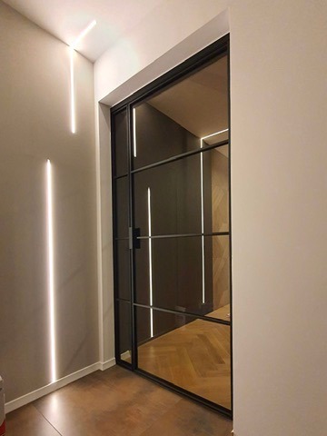 Przesuwne drzwi z czarną ramą i szklanym wypełnieniem, z widocznym odbiciem parkietu jodełkowego i oświetlenia LED na ścianie, w minimalistycznym wnętrzu.