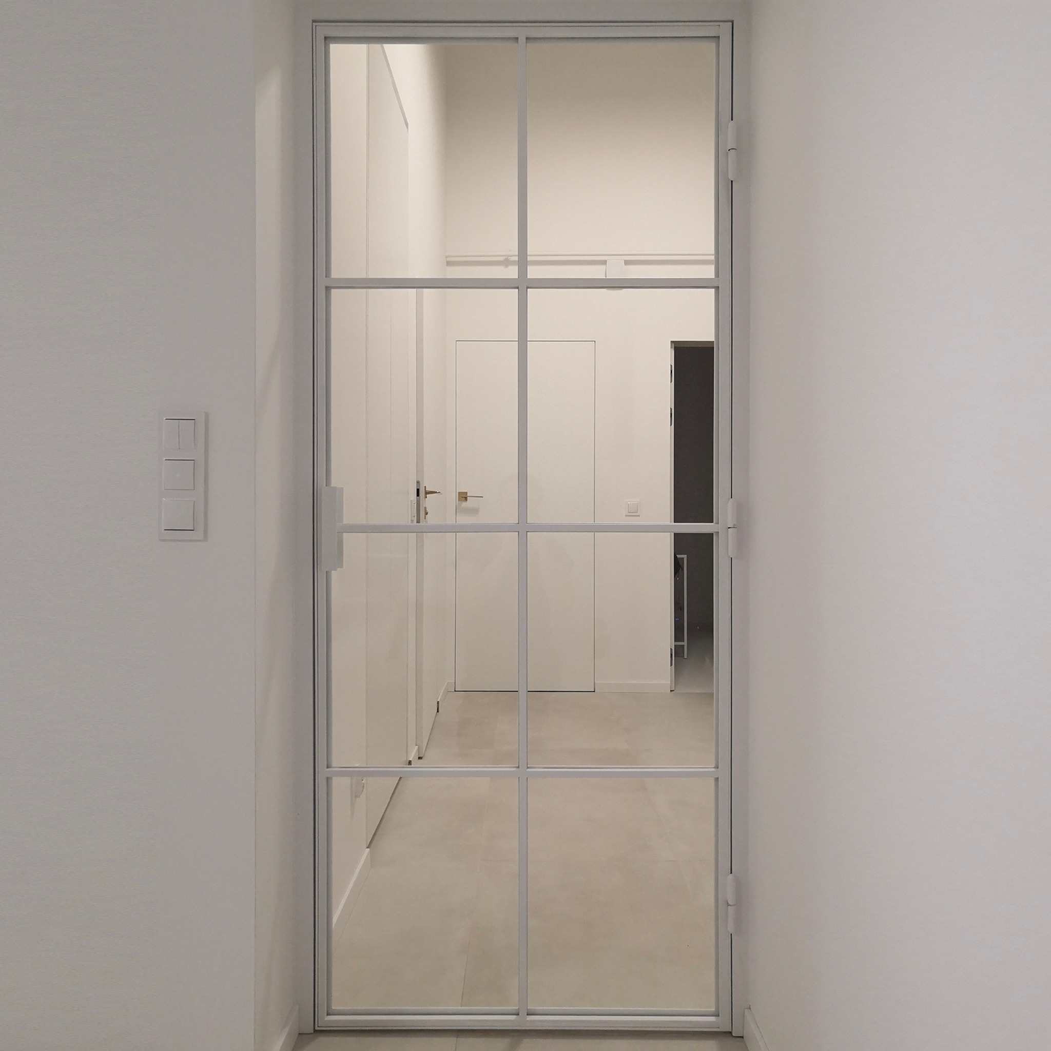 Białe, szklane drzwi z podziałami na prostokąty, prowadzące do jasnego korytarza z białymi drzwiami i szafami w tle. Po lewej stronie ściany widoczny panel z trzema włącznikami światła.