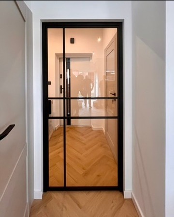 Nowoczesne drzwi z czarną ramą i szklanymi panelami, widok z korytarza z parkietem ułożonym w jodełkę, odbicie sylwetek w szkle.