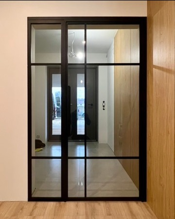 Nowoczesne, czarne drzwi z przeszkleniami w stylu loft, widok na przedpokój z drewnianą szafą i zewnętrznymi drzwiami.