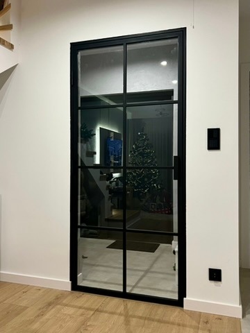 Nowoczesne drzwi z czarną ramą i szklanymi panelami, widoczne z perspektywy wnętrza domu, z choinką świąteczną odbijającą się w szkle.