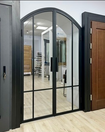 Dwuskrzydłowe, łukowe drzwi z czarnymi szprosami i przezroczystym szkłem, prezentowane w salonie obok innych modeli drzwi.