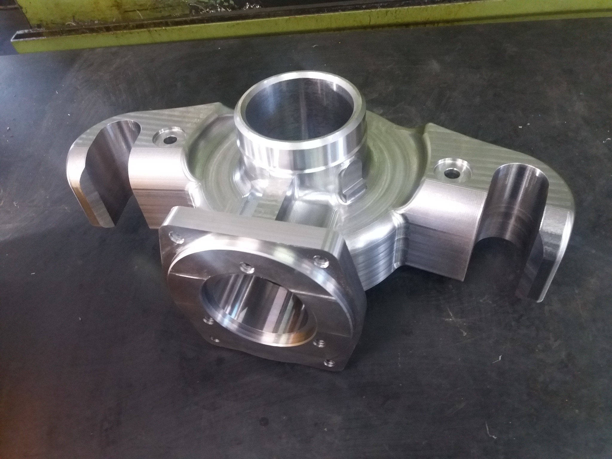 Precyzyjnie obrobiony, aluminiowy element z widocznymi śladami frezowania, leżący na ciemnym, metalowym stole warsztatowym. Element posiada centralny otwór cylindryczny i boczne mocowania...