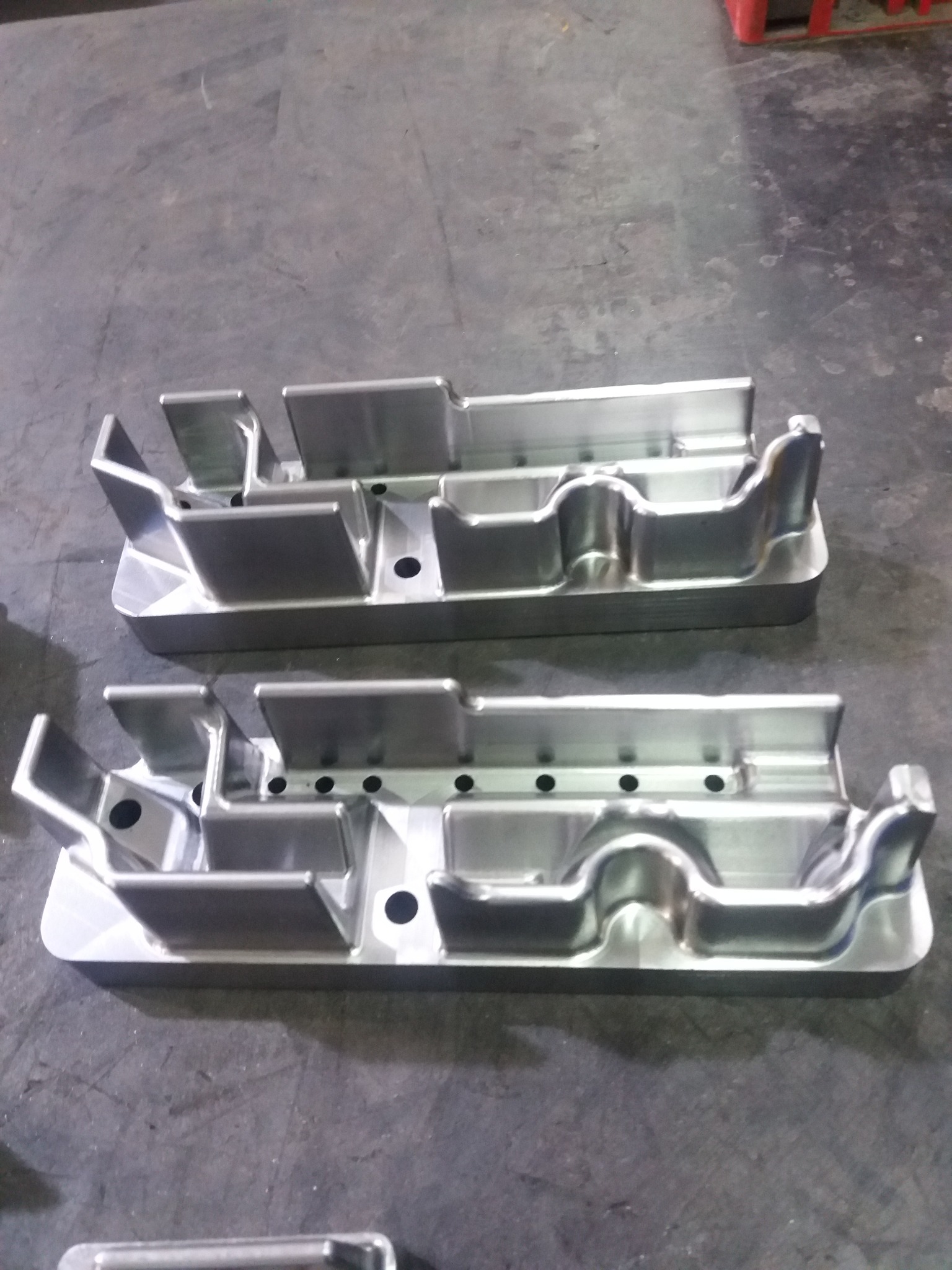 Dwa identyczne, precyzyjnie obrobione metalowe elementy z otworami, leżące na szarej, matowej powierzchni roboczej, ukazujące detale frezowania i wykończenia.