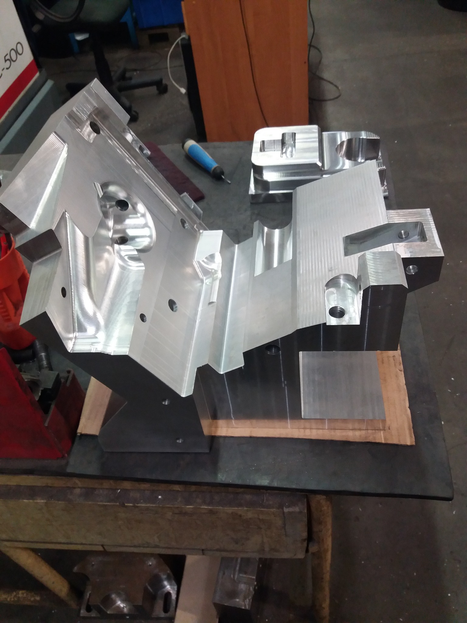 Precyzyjnie obrobiony, srebrny blok metalu z licznymi otworami i frezowaniami, leżący na stole w warsztacie. Widoczne narzędzia i elementy obrabiane w tle.