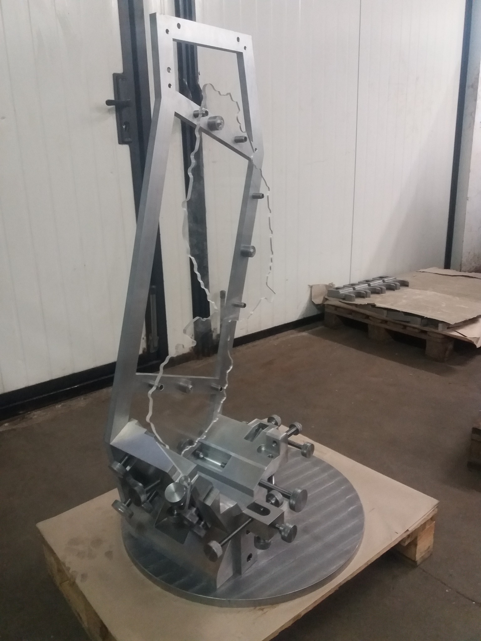 Metalowa konstrukcja z wyciętym kształtem, prawdopodobnie element maszyny, na palecie w hali produkcyjnej. Widoczne ślady obróbki.