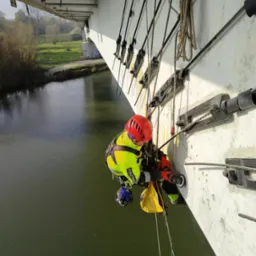Pracownik w kasku i odzieży ochronnej, wiszący na linach pod mostem nad rzeką, szlifuje betonową konstrukcję za pomocą szlifierki kątowej.