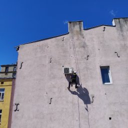 Sierra Prace Wysokościowe - Pracownik w uprzęży, na linie, montuje klimatyzator na elewacji budynku. Widok z dołu, błękitne niebo w tle. Charakterystyczne zdobienia elewacji.