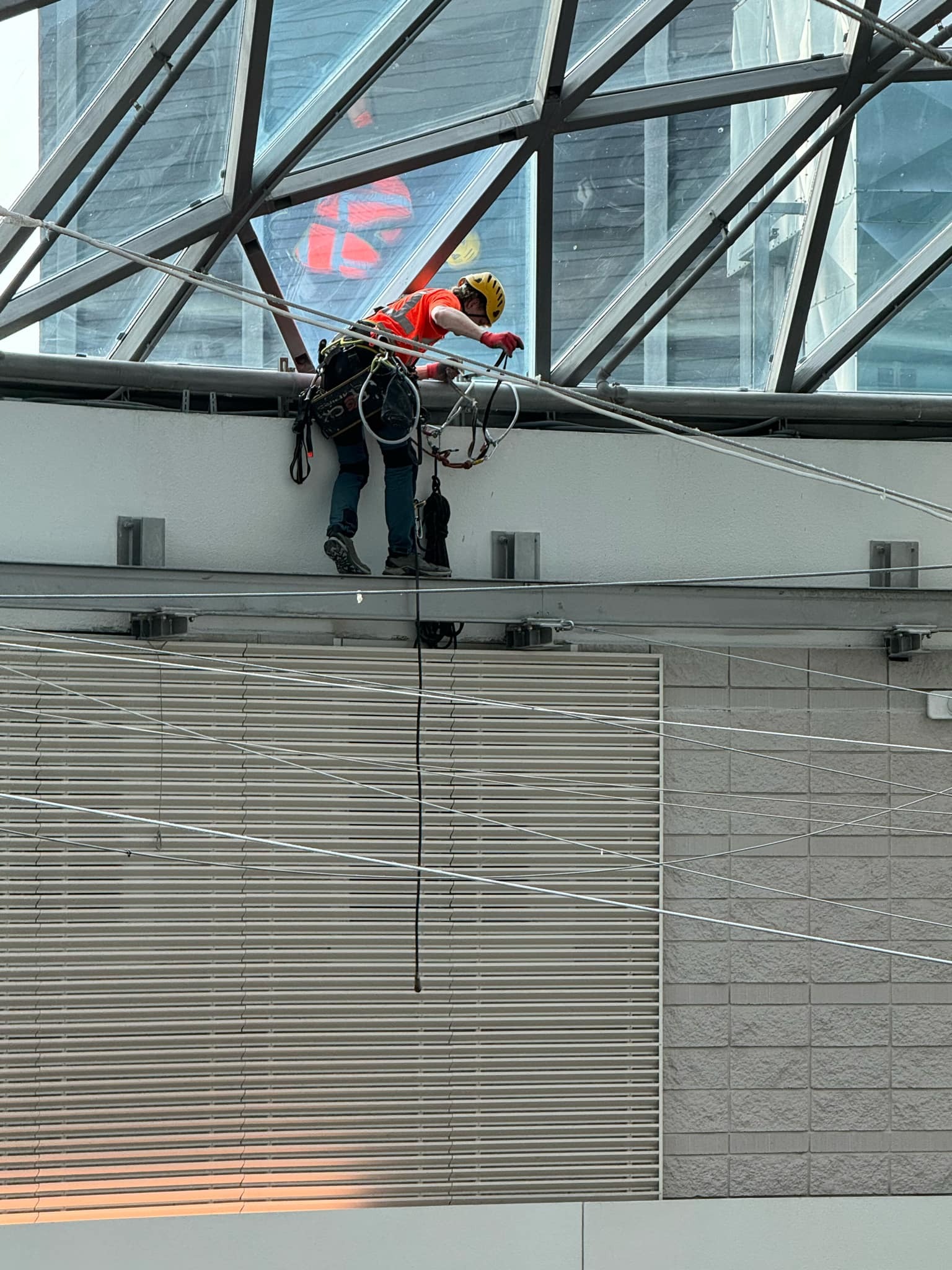 Pracownik w kasku i uprzęży, zabezpieczony linami, wykonuje zadanie na wysokości wewnątrz przeszklonej konstrukcji, prawdopodobnie w atrium budynku.