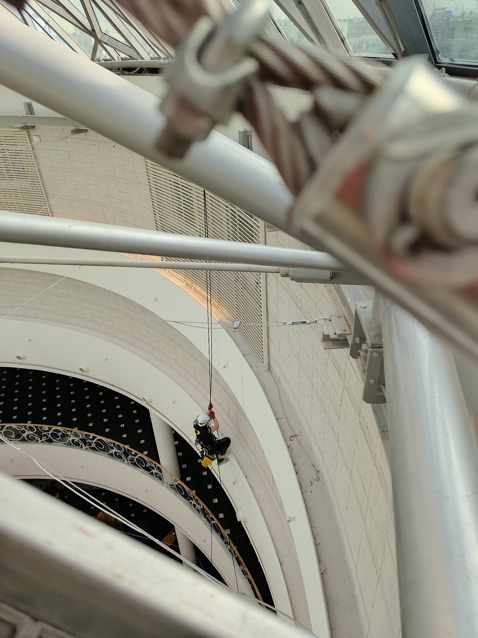 Pracownik na linach wewnątrz budynku z przeszklonym dachem, widok z góry na atrium z balustradami.